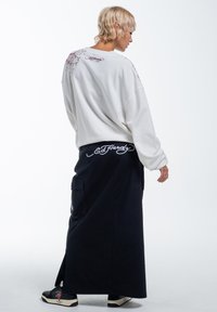 Hvid sweatshirts med broderede designs på ryggen, parret med en lang sort nederdel med cargo-lommer og hvid logo-detalje. Sorte sneakers.