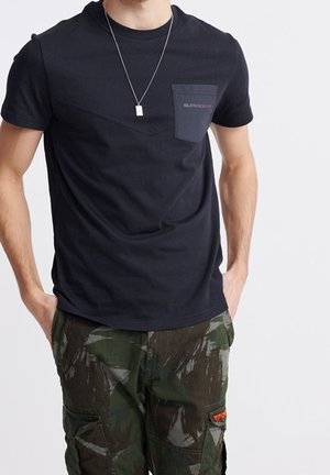 Czarny t-shirt z krótkim rękawem z kieszonką na piersi i kontrastowym trójkątnym panelem; w połączeniu z zielonymi spodniami cargo w kamuflażu z bocznymi kieszeniami.