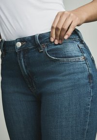 Jeans en denim bleu foncé avec une coupe slim, un design classique à cinq poches, des rivets en cuivre et un bouton argenté à la taille.