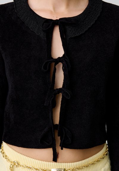 Cárdigan corto negro hecho de una suave tela texturizada. Cuenta con un cuello en forma de concha, un cierre frontal atado y detalles de lazo intrincados.