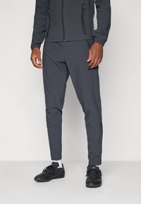 Pantalons de sport gris à coupe ajustée, dotés de poches zippées et de poignets élastiques. Associés à des chaussures noires et des chaussettes blanches.