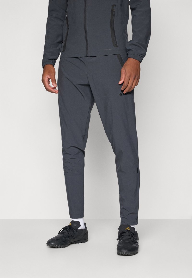 Pantalons de sport gris à coupe ajustée, dotés de poches zippées et de poignets élastiques. Associés à des chaussures noires et des chaussettes blanches.