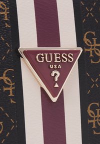 Złote logo Triangular Guess USA na burgundowym tle, z białą i czarną paskowaną teksturowaną powierzchnią w tle.