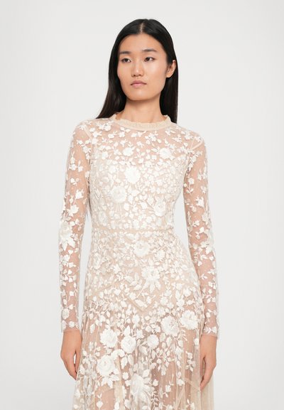 Vrouw in een lichtbeige lange mouwen doorschijnende jurk met witte bloemenborduursels, staande tegen een effen lichte achtergrond.
