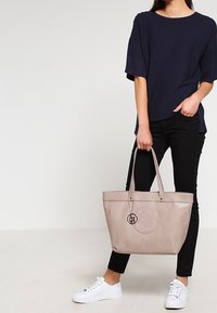 Borsa a tote in pelle beige con logo in rilievo, due manici lunghi e charm in metallo. Indossata con pantaloni neri e sneakers bianche; top a trama.