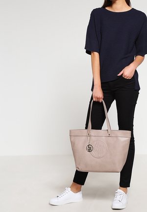 Beżowa skórzana torba na ramię z wytłoczonym logo, dwoma długimi uchwytami i metalowym charms'em. Noszona z czarnymi spodniami i białymi sneakersami; teksturowana bluzka.