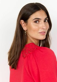 Rotes Oberteil mit Spitzenverzierungen an den Schultern, Rundhalsausschnitt und kurzen Ärmeln. Das Modell hat lange, glatte braune Haare und trägt goldene Ohrringe.