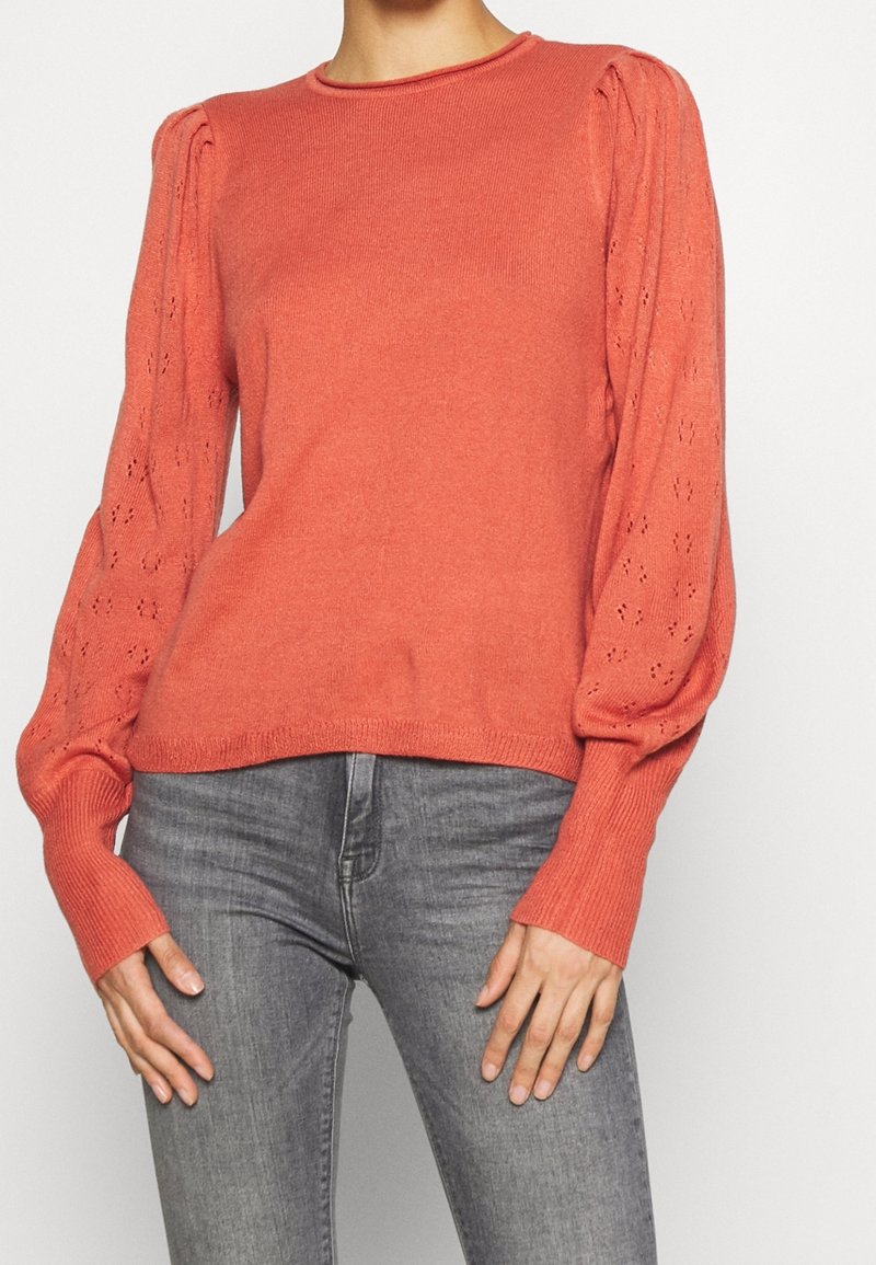 Femme portant un pull en maille corail à manches longues avec des manches bouffantes à motifs ajourés et un jean gris ajusté, debout devant un fond uni.