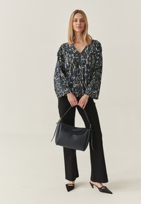 Langärmlige Bluse mit blauem, beigem und schwarzem abstraktem Muster; schwarze Schlaghose; eine dunkelblaue strukturierte Handtasche; schwarze spitze Schuhe.