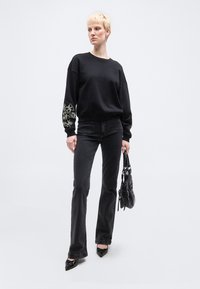 Γυναίκα με κοντά ξανθά μαλλιά που φορά μαύρο φούτερ με κέντημα στους αγκώνες, μαύρα τζιν, μυτερά μαύρα παπούτσια, κρατώντας μαύρη τσάντα.