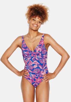 Donna con capelli ricci sorridente, che indossa un costume intero floreale blu e rosa, con le mani sui fianchi, su uno sfondo bianco uniforme.