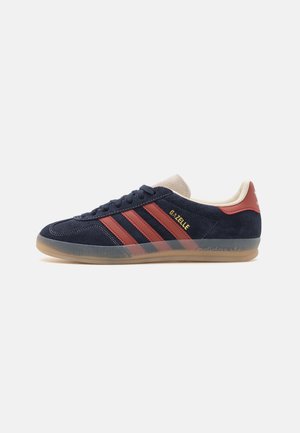Low-profile Adidas Gazelle sneaker in navy suede with rust red stripes, beige tongue, and translucent gum sole, side view.