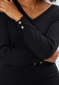 Schwarzer Strickpullover mit V-Ausschnitt, langen Ärmeln mit dekorativen goldenen Knöpfen und körperbetontem Schnitt. Weicher Stoff, klassisches Design.