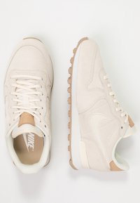 Beiga mockaskor med krämfärgade detaljer, som har en vit mellansula och en brun gummisula, snörning och logotyp på tungan.