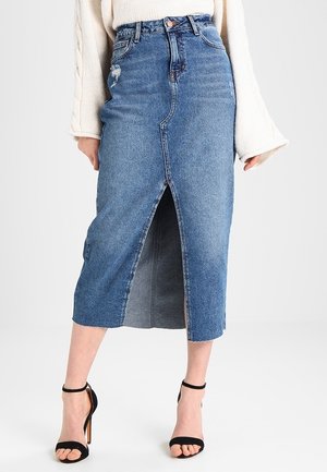 Femme portant une jupe midi en denim bleu taille haute avec une fente sur le devant, associée à des talons hauts noirs à bride cheville et un pull blanc.