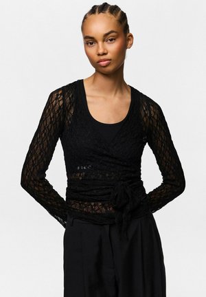 Femme aux cheveux tressés portant un top cache-cœur en dentelle noire à manches longues sur un débardeur noir et un pantalon noir taille haute, debout les mains dans le dos.