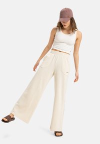 Cream-colored wide-leg pants with two front pockets, paired with a white crop tank top and a light mauve baseball cap. Brown slide sandals.