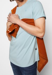 Ljusblå bomullströja med korta ärmar, tillsammans med en orange manchesterjacka knuten runt kroppen. Jeans synliga från midjan och ner.