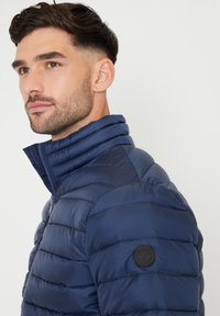 Giacca imbottita blu navy con pannelli cuciti, colletto alto e una toppa rotonda sul braccio sinistro. Materiale liscio e leggero.