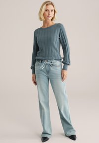 Maglione a coste blu con maniche lunghe, abbinato a jeans a gamba dritta di un blu chiaro. Gli accessori includono una cintura e scarpe nere con punta.