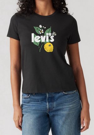 Femme portant un t-shirt Levi's noir avec un logo blanc, des feuilles vertes, des fleurs blanches et un motif de citron jaune, associé à un jean bleu.
