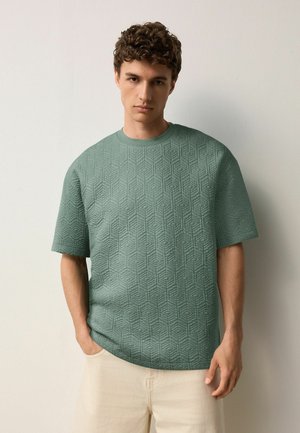 Grøn, struktureret t-shirt med et geometrisk mønster; korte ærmer, rund hals, afslappet pasform; båret med lyse bukser.