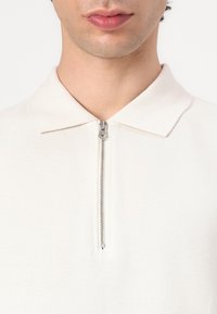White polo shirt with a zip closure, made from textured fabric. Features a flat collar and short sleeves, showcasing a clean, minimalist design.