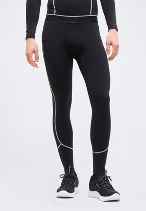 Moški v črnih oprijetih športnih legicah z belim šivom in črnih tekaških čevljih z belimi podplati, stoji na enobarvnem ozadju.