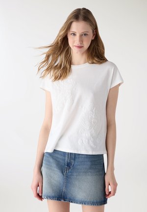 Giovane donna con capelli biondi mossi indossa una maglietta bianca a maniche corte ricamata e una mini gonna di jeans sbiadita su uno sfondo semplice.