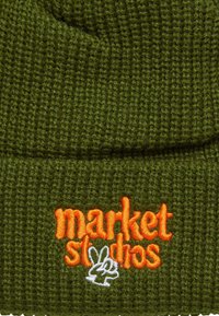 Zelená pletená čiapka s textúrovaným povrchom. Obsahuje oranžový vyšitý nápis "market" a malé biele logo zajaca.