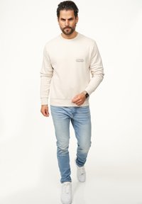 Helles beiges Sweatshirt mit Rundhalsausschnitt und gerippten Bündchen, mit einem kleinen Grafikdruck auf der Brust. Kombiniert mit hellblauen Jeans und weißen Sneakers.