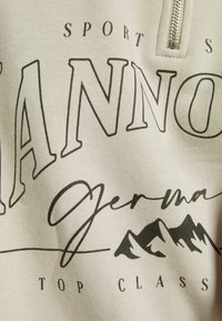 Beige sweatshirt med en fjärdedelsdragkedja. Har svart tryckt text och berggrafik. Mjuk textur, avslappnad passform, avslappnad design.
