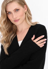Cardigan nero a maglia con scollatura a V, materiale a coste texturizzato e maniche lunghe. Il modello indossa una maglietta bianca sotto.