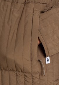 Brun, vadderat yttertyg med vertikal söm och sidoficka. Har en dragkedjsöppning och en etikett med texten "RAINS".