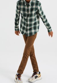 Zeleno-bílá kostkovaná košile na knoflíky, hnědé kalhoty a vícebarevné tenisky s černými a bílými akcenty. Dlouhé rukávy, volný střih.