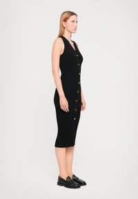 Robe midi noire à côtes avec un décolleté en V profond, comportant des accents de boutons sur le devant. Le mannequin porte des mocassins noirs. Texture lisse.
