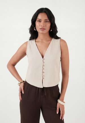 Woman with wavy black hair wears sleeveless beige buttoned top, dark brown pants, gold hoop earrings, and chunky bracelets against white background.