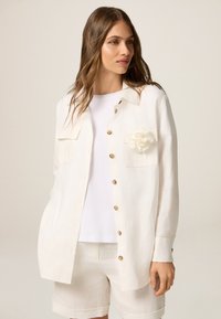 Camicia bianca in lino con maniche lunghe, tasca sul petto e dettaglio di un fiore in tessuto. Dispone di chiusura a bottone e vestibilità comoda. Abbinata a pantaloni corti bianchi.
