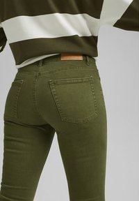 Jeans skinny vert olive avec une texture lisse, présentant une étiquette en cuir à l'arrière et deux poches arrière, montrés sous un angle bas.