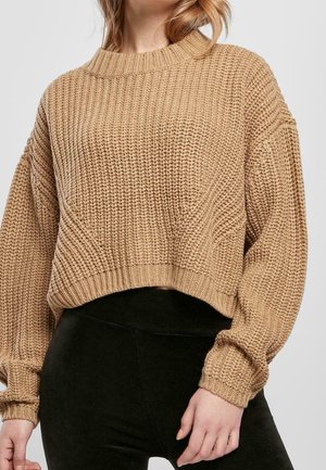 Tułów kobiety ubranej w beżowy, gruby sweter z bufiastymi rękawami, zestawiony z czarnymi, wysokimi spodniami na szarym tle.