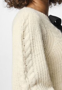 Κρεμ πλεκτό πουλόβερ με σχέδιο από καλώδιο στα μανίκια, με απαλή υφή και μαύρη κορδέλα στη λαιμόκοψη.