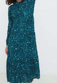 Abito verde acqua con design a strati e chiusura con bottoni, caratterizzato da una stampa leopardata bold in nero e blu su un tessuto leggero.