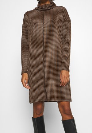 Femme portant une robe à manches longues marron et noire à motif pied-de-poule avec un col bénitier et des bottes noires montant jusqu'aux genoux.