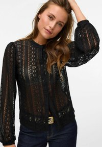 Mustmustest lace-pluus, millel on keerukad mustrid, pikad puff-sleevid, nööpidega sulgemine ja läbipaistev tekstuur. Sobitatud tumedate teksade ja vööga.
