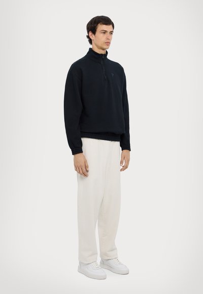 Felpa nera mezza zip con orlo e polsini a costine, abbinata a pantaloni bianchi a gamba larga e sneaker bianche. Design semplice, look moderno.