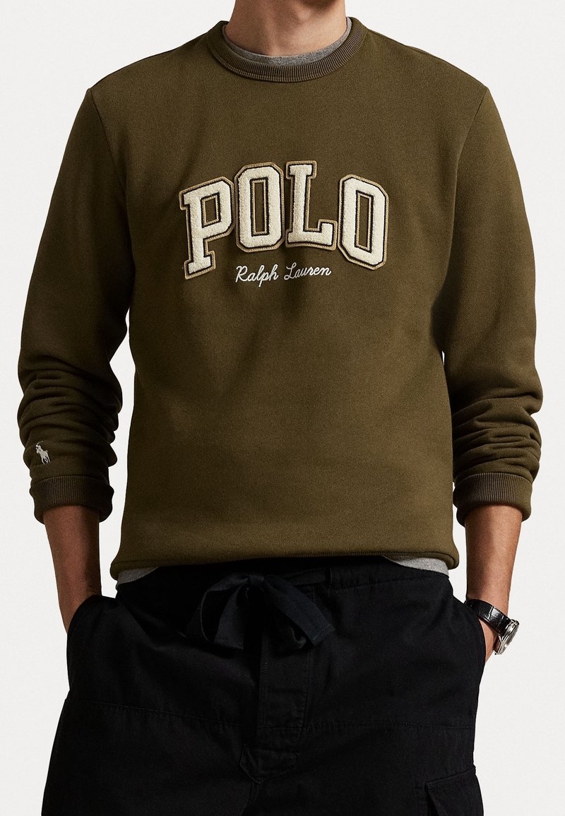 Homme portant un sweat-shirt olive à col ras du cou avec le texte "POLO Ralph Lauren" et un pantalon noir, les mains dans les poches.
