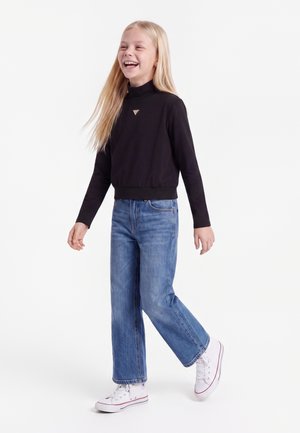 Ragazza sorridente con lunghi capelli biondi cammina indossando una maglia nera a maniche lunghe, jeans blu e scarpe da ginnastica bianche su uno sfondo bianco uniforme.