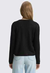 Persoon met lang blond haar die een zwarte trui met lange mouwen en lichtblauwe spijkerbroek draagt, van achteren gezien tegen een lichte achtergrond.