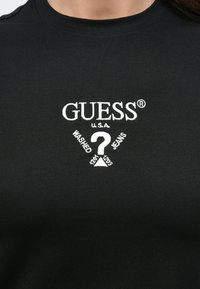 Μαύρο βαμβακερό T-shirt με λευκό κεντητό λογότυπο "GUESS" και σχέδιο με ερωτηματικό, με το κείμενο "WASHED JEANS" από κάτω, σε στυλ με στρογγυλή λαιμόκοψη.