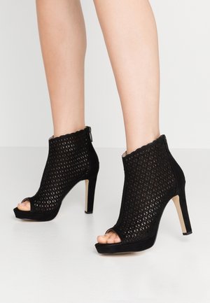 Czarne sandały na wysokim obcasie z odkrytym palcem, wykonane z miękkiego zamszu o teksturowanym, wyciętym wzorze. Posiadają faliste krawędzie oraz boczny zamek błyskawiczny.